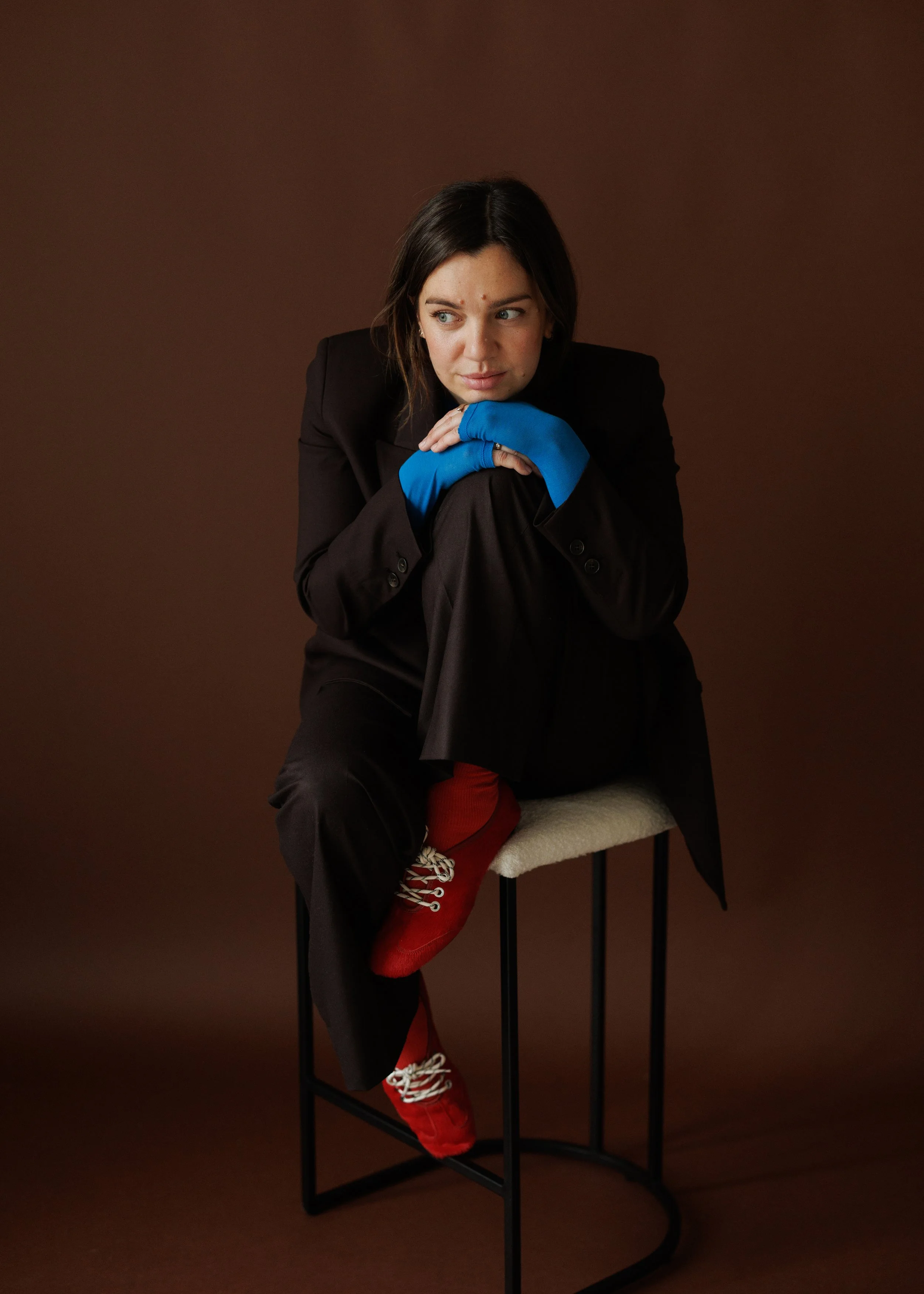 A portrait of Lottie Topping, Wearing a brown blazer, blue gloves, red shoes, and red socks, sitting on a high stool against a plain brown background, with her arms resting on her knees and looking to the side.