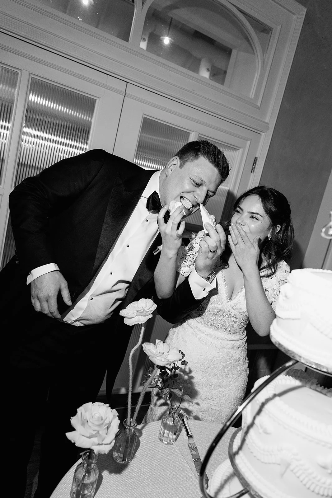 A bride and groom at a wedding reception, with the groom taking a bite of a piece of wedding cake and the bride covering her mouth, both smiling.