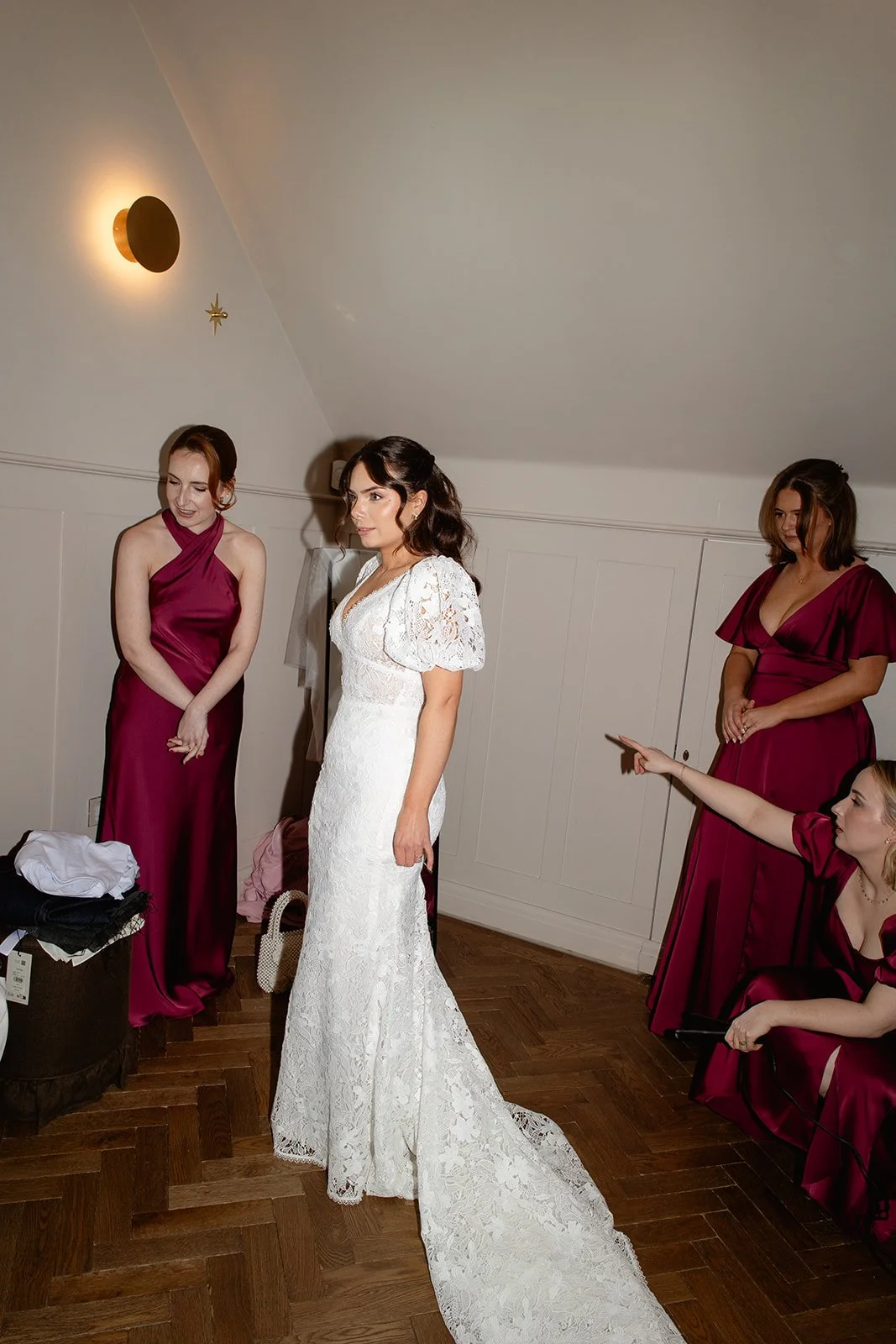 Bridal party with one woman in a white lace wedding dress, three women in matching maroon dresses, in a room with wood flooring and soft lighting.
