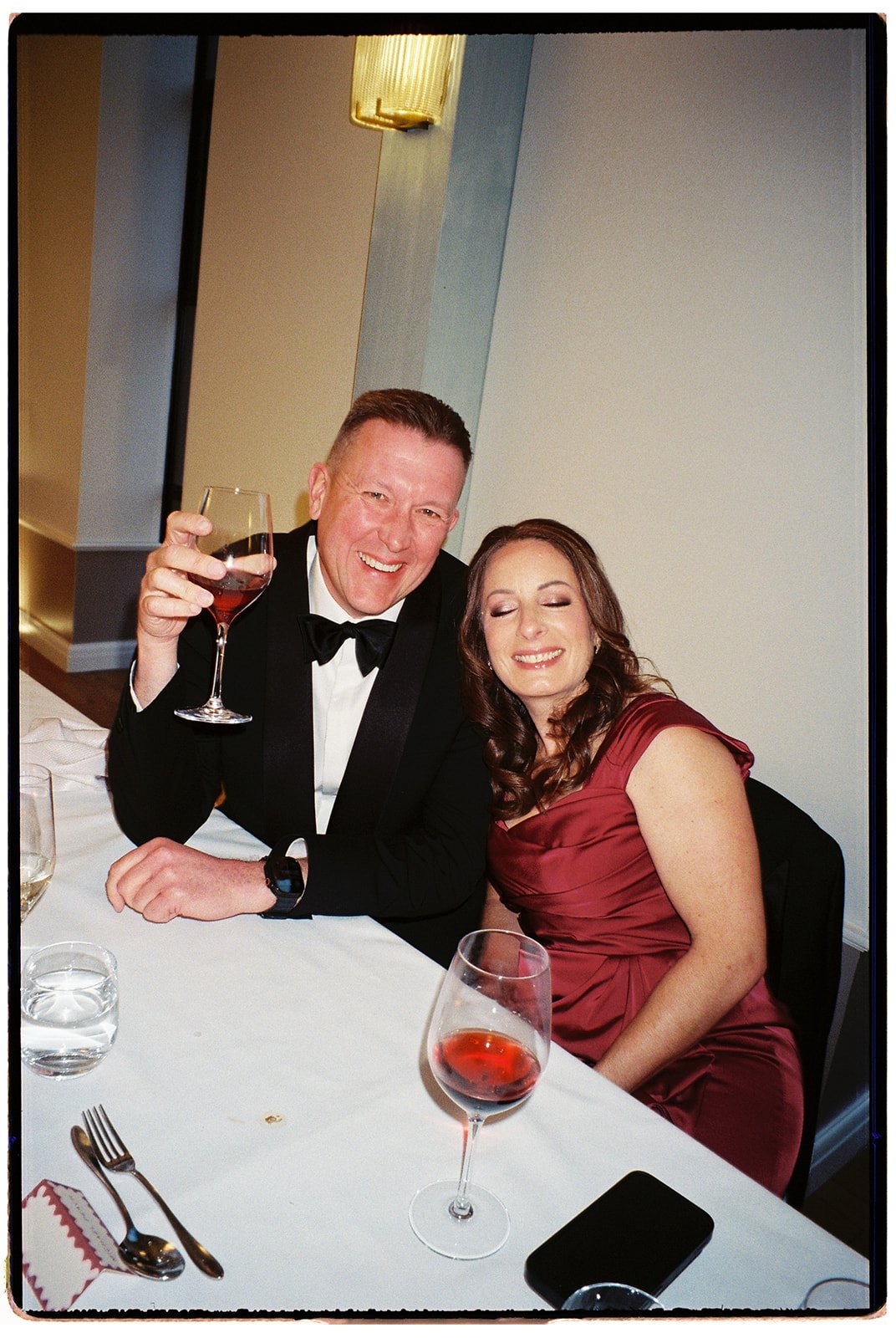 A man in a tuxedo and a woman in a red dress sitting at a dinner table, smiling and celebrating with wine glasses at a formal event.