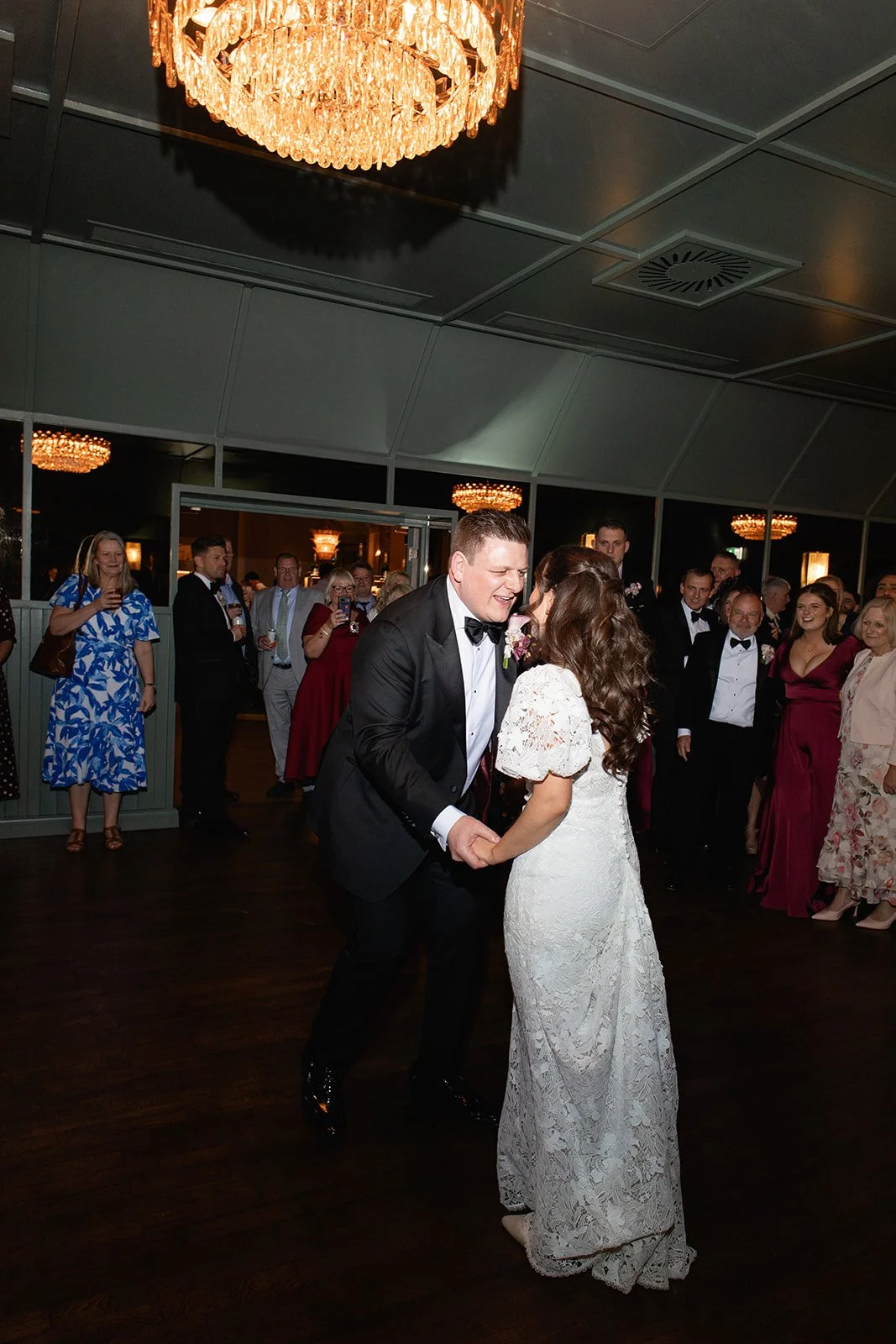 A bride and groom dancing at their wedding reception with guests watching and smiling in the background, indoors with chandeliers overhead.