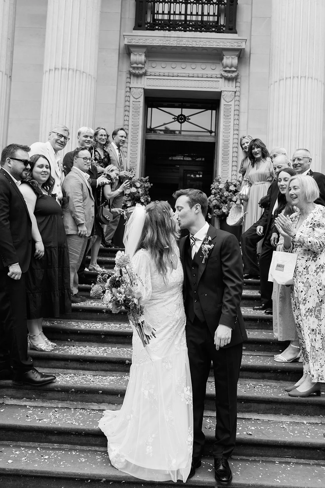 Black and white photo of a newlywed couple kissing on the steps of a building, surrounded by friends and family celebrating their wedding.