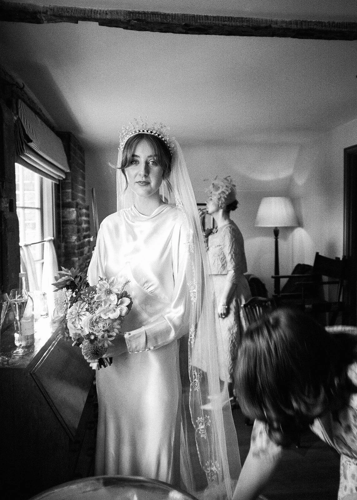 A bride in a satin dress with a veil and crown, holding a bouquet, standing indoors near a window. A woman in a headscarf and a person with dark hair are nearby, in a room with a lamp and chairs.