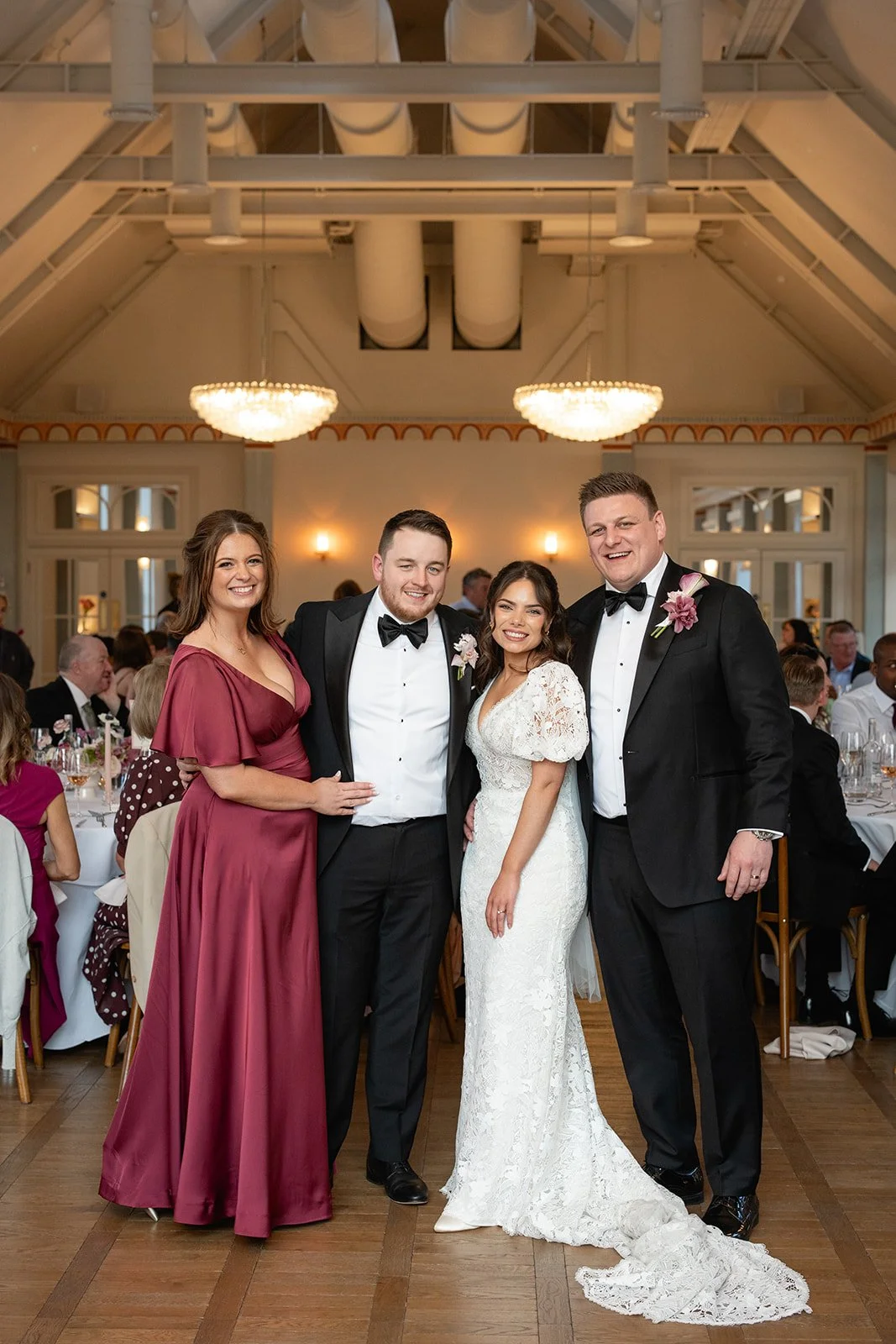 A group of four people dressed in formal wedding attire standing together at a wedding reception. The woman in center is wearing a white lace wedding dress, and the two men are in black tuxedos with bow ties. The woman on the left is wearing a maroon