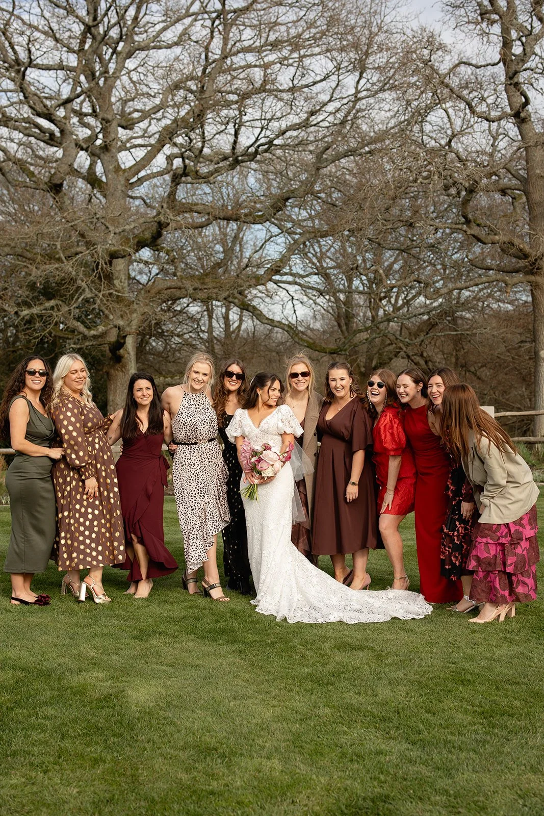 A group of women, including a bride in a white wedding dress holding a bouquet, standing outdoors on a grassy area with leafless trees in the background, all smiling and posing for a photo.