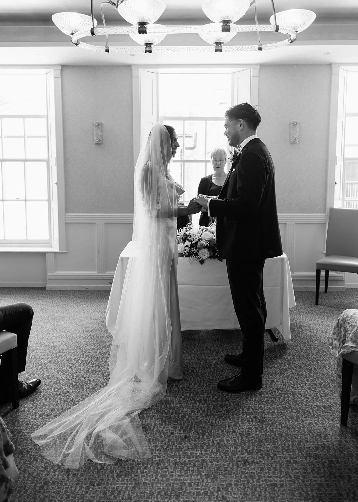 A black-and-white photo of a wedding ceremony with a bride and groom holding hands and smiling at each other, an officiant behind them, in a bright room with large windows and a chandelier.