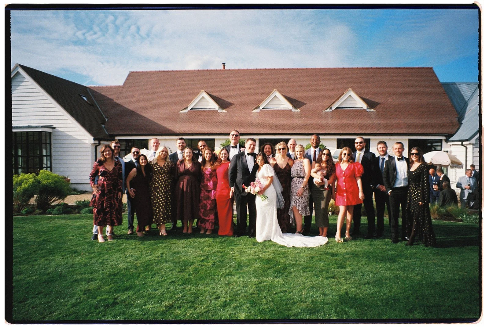 A large group of people dressed in formal attire, including a bride in a white wedding dress holding a bouquet, and a groom in a black tuxedo, standing outdoors on a grassy area in front of a house with a brown roof and white siding, on a sunny day.