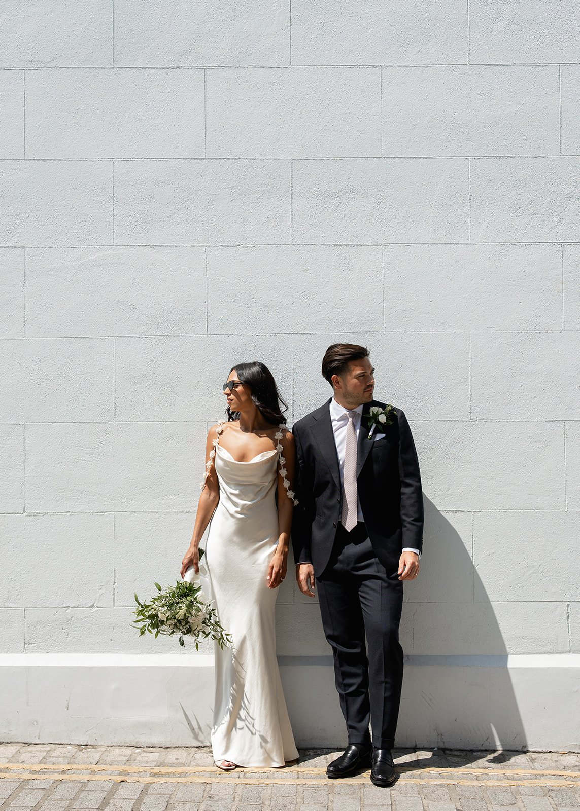 A bride and groom standing against a white wall on their wedding day. The bride is wearing a white wedding dress and holding a bouquet, while the groom is in a black suit with a white shirt and tie, both looking in different directions.