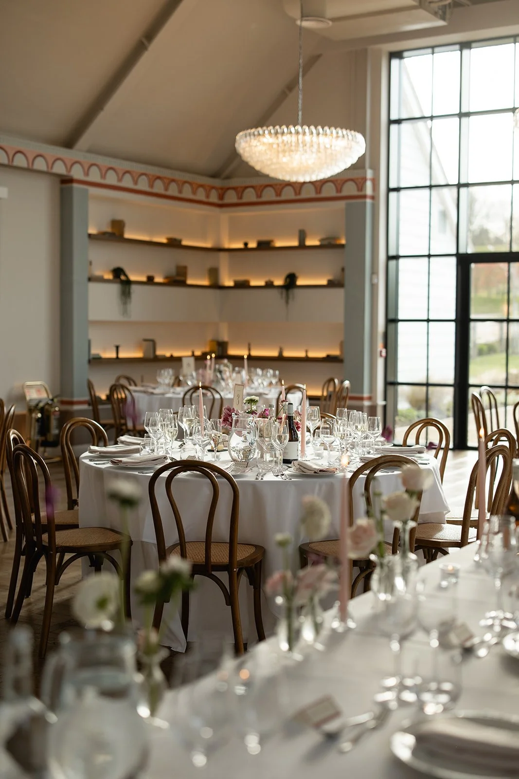 Elegant dining room with round table set for a formal event, decorated with flowers, candles, wine, and glassware, large window with outside view, and modern shelving with decor.