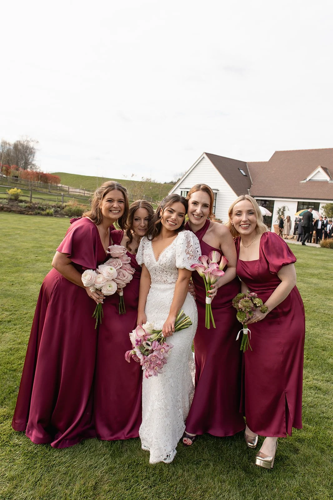 A bride and four bridesmaids posing outdoors on a lawn during a wedding celebration, holding bouquets of flowers.