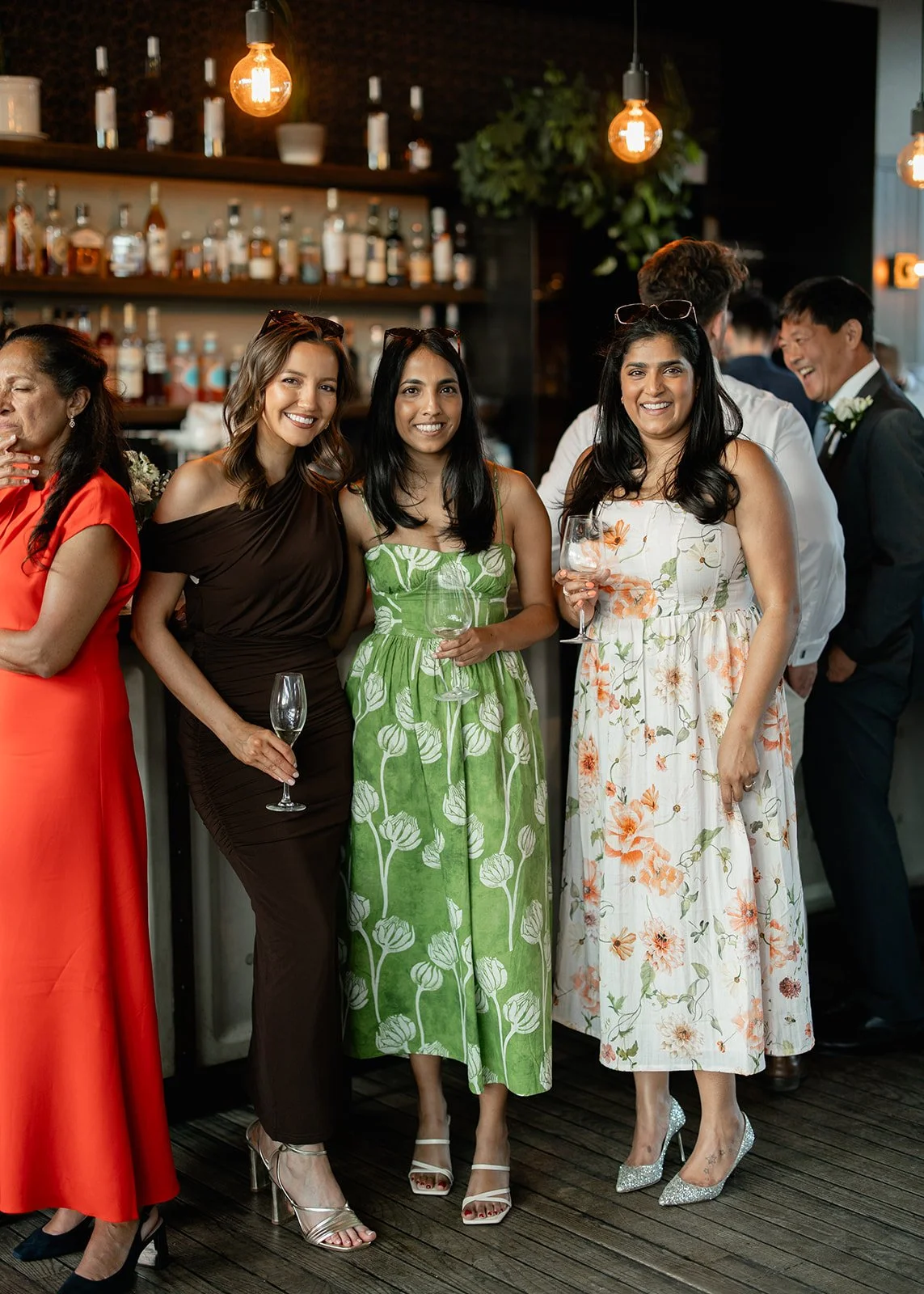 Group of women in colorful dresses holding wine glasses at a social event in a bar or restaurant, with a bar and bottles in the background.