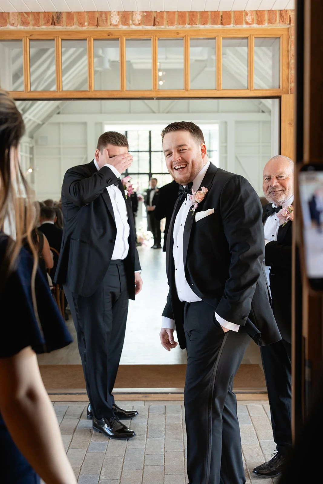 A group of men in tuxedos at a wedding reception, smiling and laughing, with one man covering his eyes and another man standing confidently with his hand in his pocket.