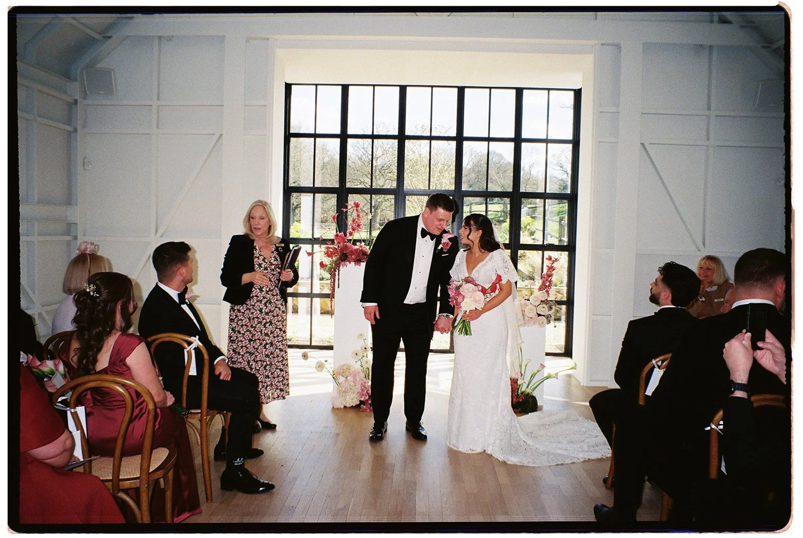 A wedding ceremony indoors with a bride and groom standing together, holding hands. The bride is holding a bouquet, and the groom is dressed in a black tuxedo. Guests are seated on either side, and a woman is speaking at the side. Large windows with 