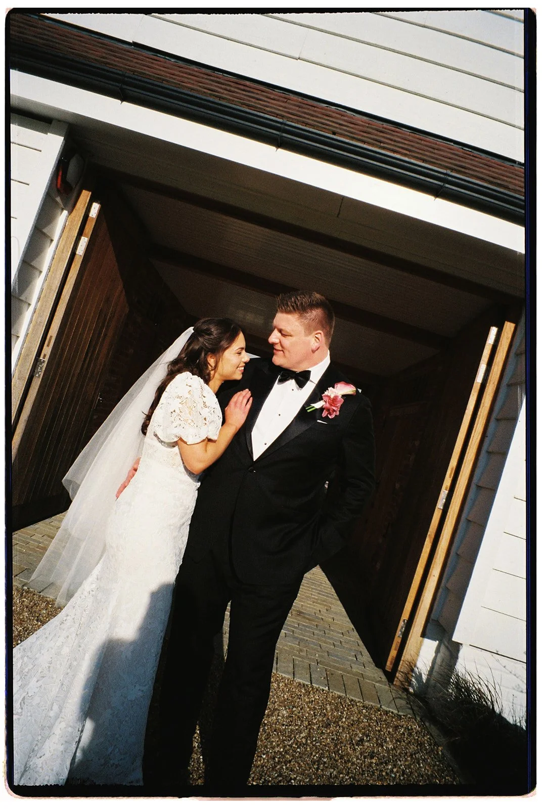 A bride in a white lace wedding dress and veil with a groom in a black tuxedo with a pink boutonniere, sharing a moment at the entrance of a building during daytime.
