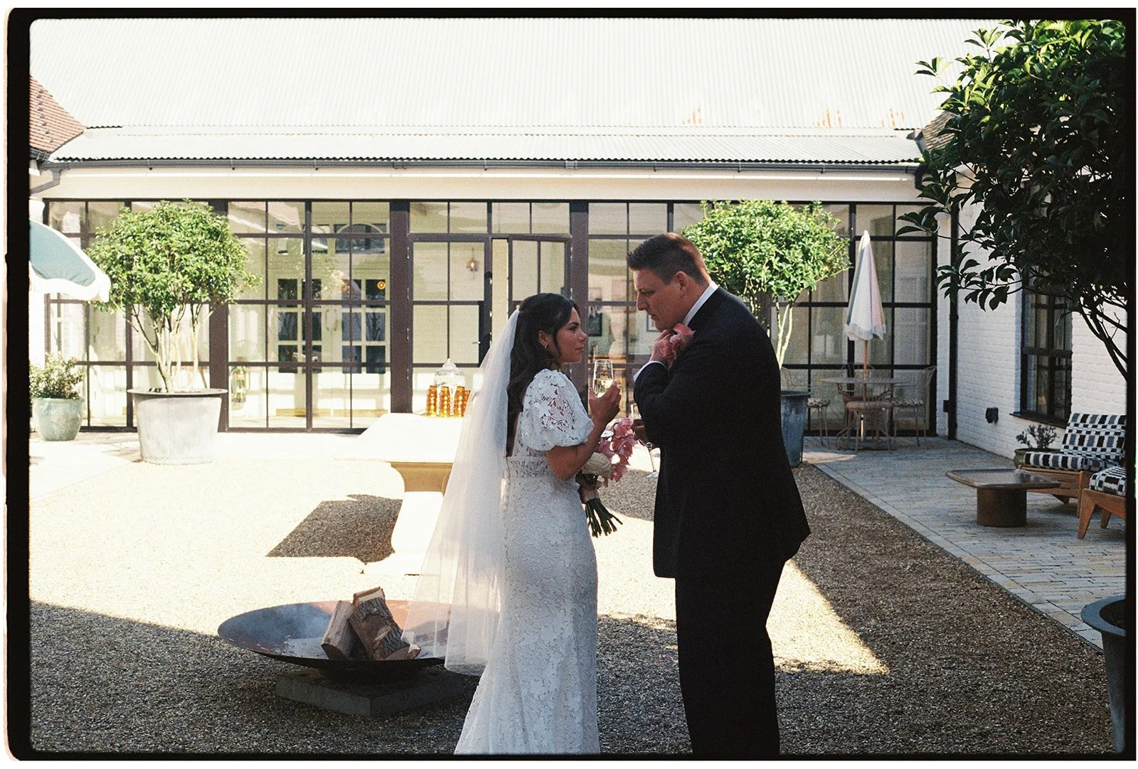 A bride and groom during their wedding ceremony outdoors, standing closely and holding champagne glasses, with a modern building and trees in the background.