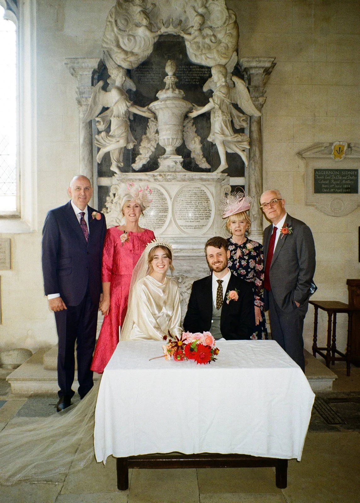 A wedding photo taken inside a church. The bride and groom are seated at a table with a bouquet of flowers. Four other people, two men and two women, are standing behind them, all dressed in formal attire. A large religious sculpture and plaque are v