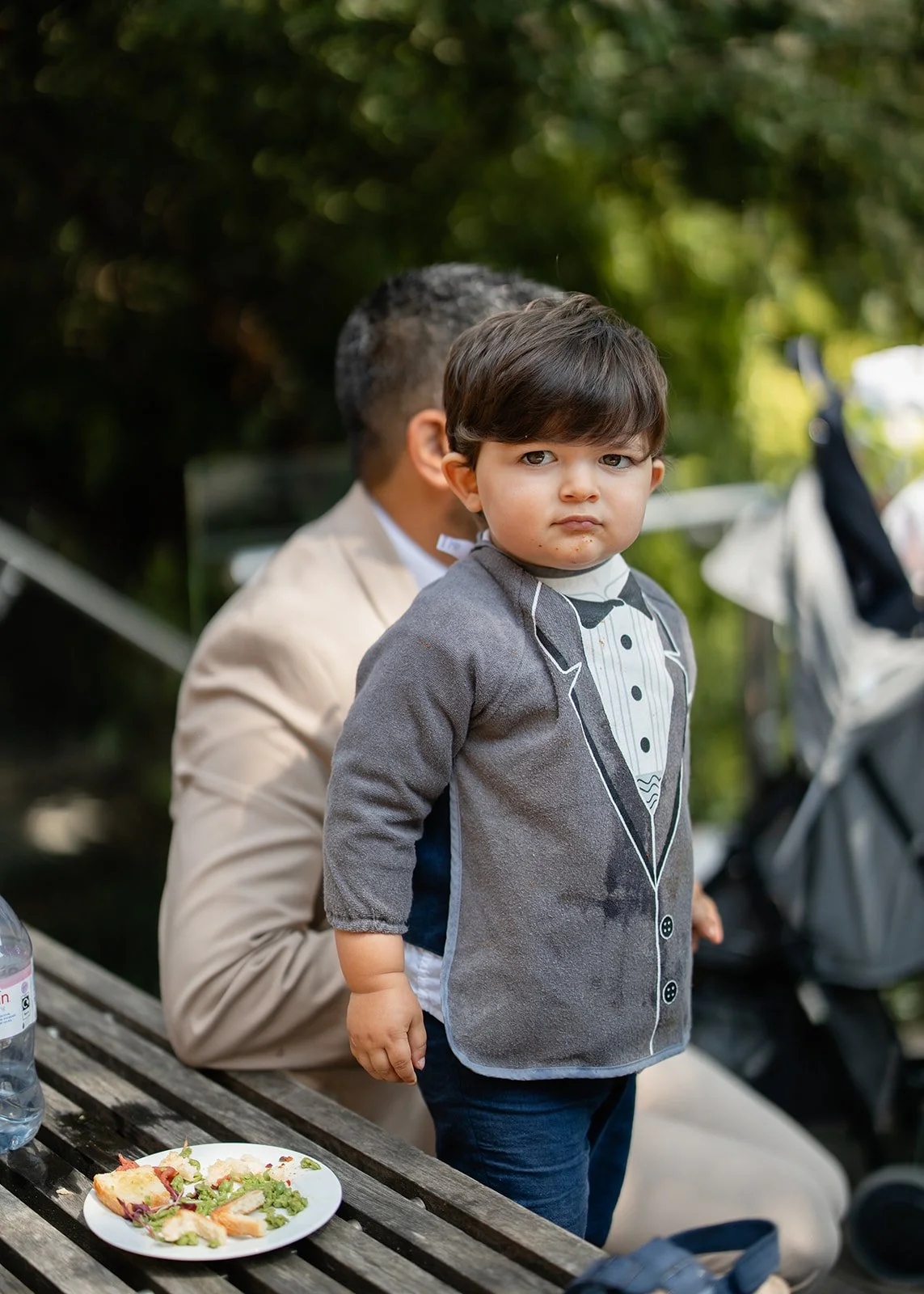 A young boy with brown hair and a serious expression standing outdoors next to a person in a beige jacket. The boy is wearing a gray jacket with a tuxedo design and dark pants. There is a plate of salad and a water bottle on a picnic table in front o