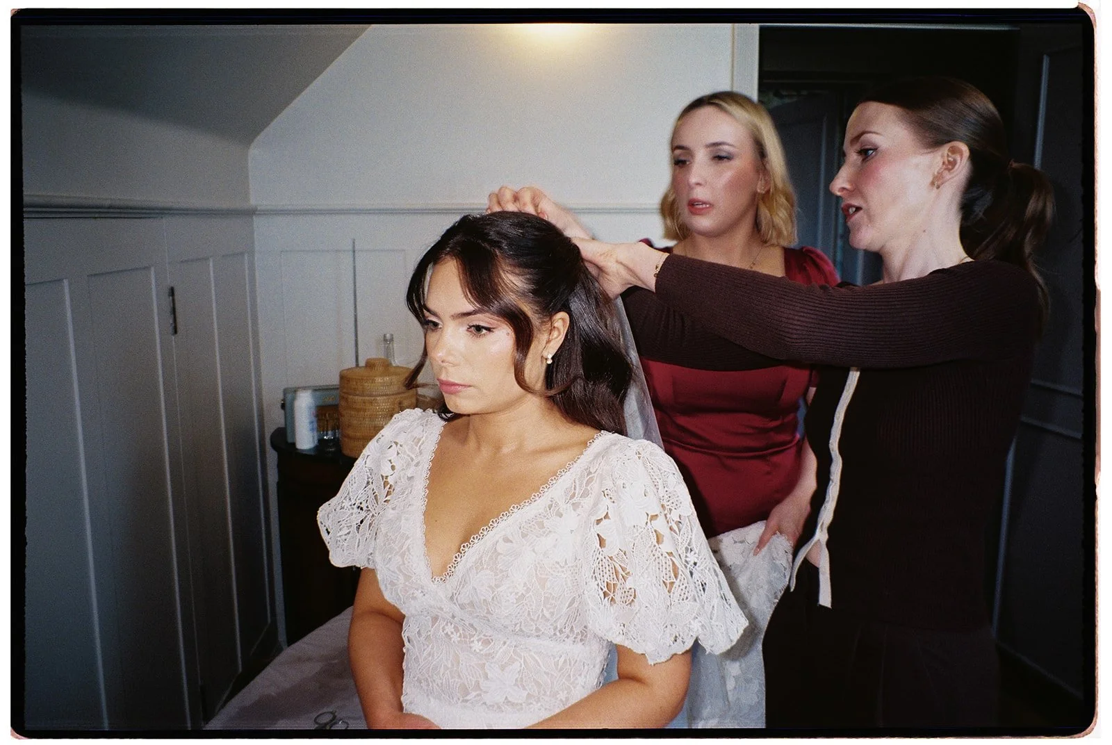 A woman in a white lace dress having her hair styled by a hairstylist while a makeup artist looks on in a dressing room.