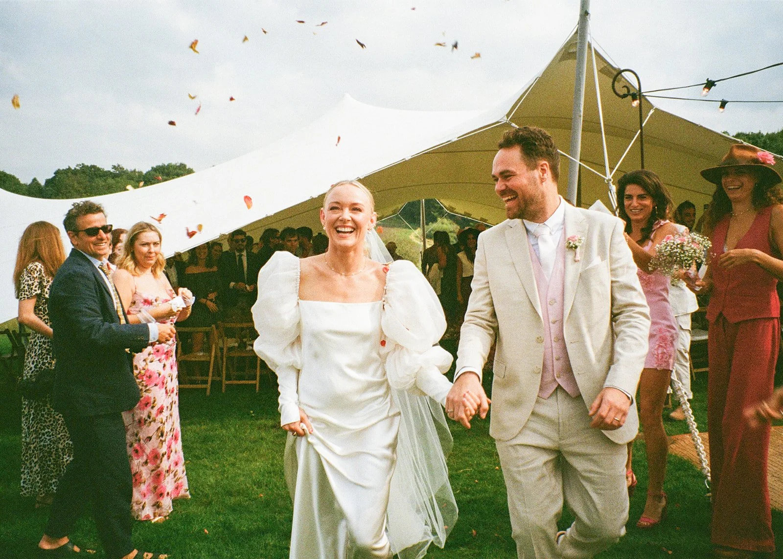 A bride and groom holding hands and smiling at each other at an outdoor wedding reception. The bride is wearing a white dress with puffy sleeves and the groom is in a light-colored suit. There are guests in colorful attire and a large white tent in the background, with confetti falling from above.