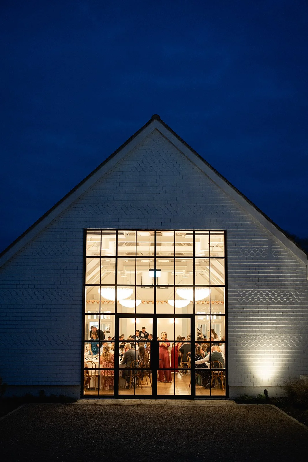 Large window showing an illuminated Poplar hall, with chandeliers and people inside at a gathering, building with white brick exterior, triangular roof, against a dark evening sky.