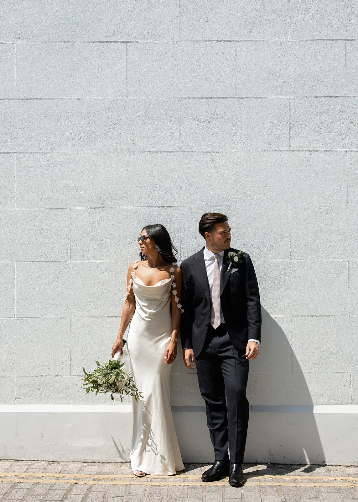 A bride and groom standing against a large white wall. The bride is wearing a white wedding dress and holding a bouquet of flowers, and the groom is dressed in a dark suit with a white shirt, tie, and boutonniere.