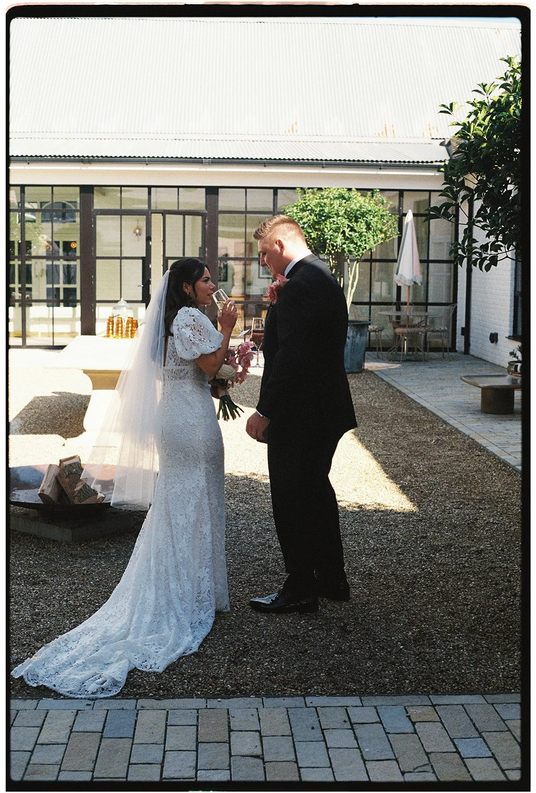 A bride and groom stand close together outdoors, holding glasses of champagne, at their wedding celebration.