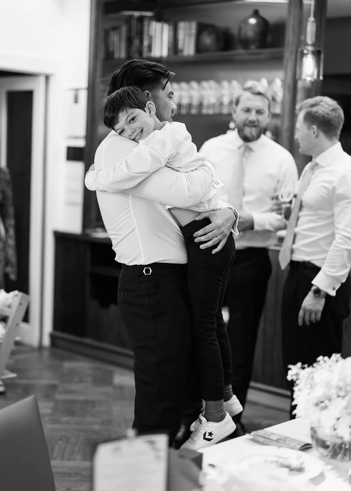 A man holding a smiling young boy in his arms, with a group of people in the background engaged in conversation, inside a warmly lit room.