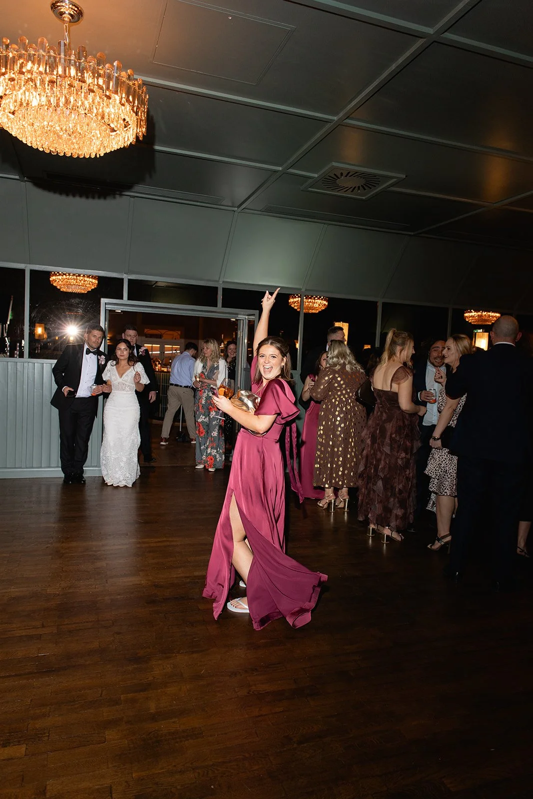 A woman in a pink gown dancing and smiling at a formal party with guests in elegant attire, a chandelier, and warm lighting.