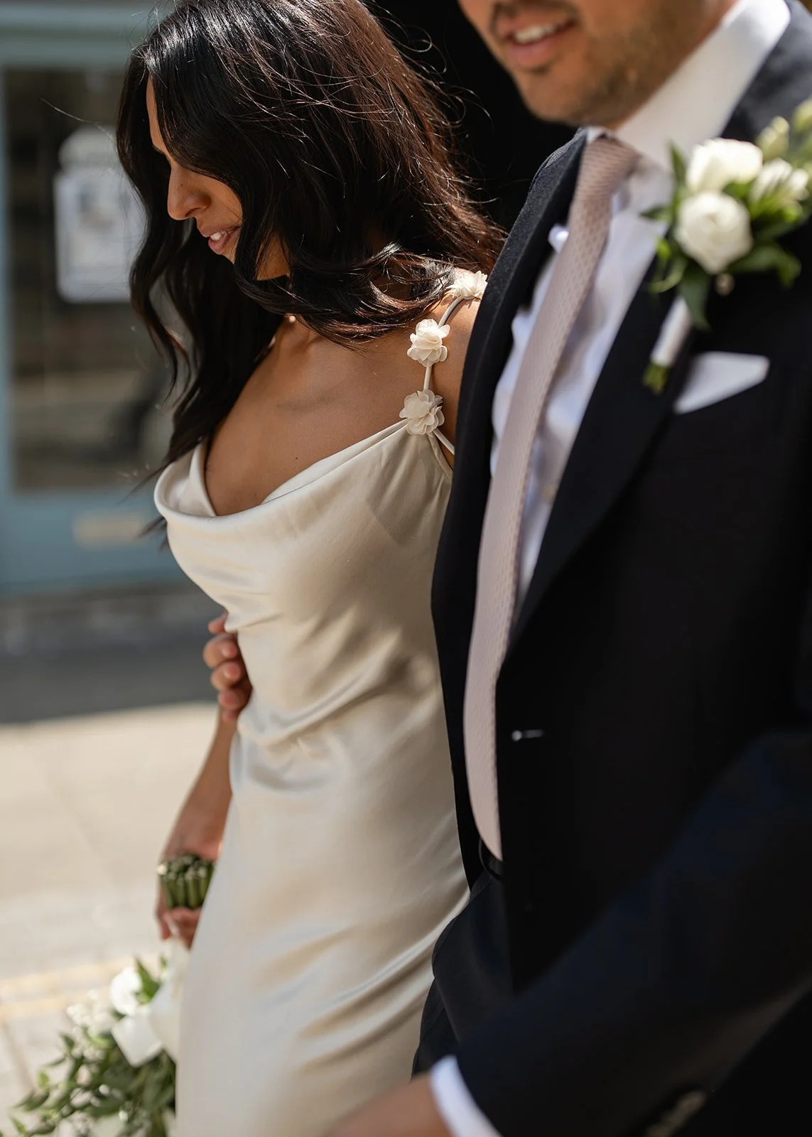 A bride and groom during their wedding ceremony, holding hands and smiling, with the bride in a cream-colored dress with floral details on her shoulder and the groom in a dark tuxedo with a white boutonniere.