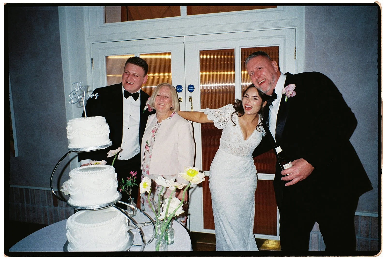 Group of five people celebrating a wedding, standing behind a multi-tiered wedding cake, with smiling faces. Two men in tuxedos, a woman in a wedding dress, and two women in formal attire, in a room with a white door in the background.
