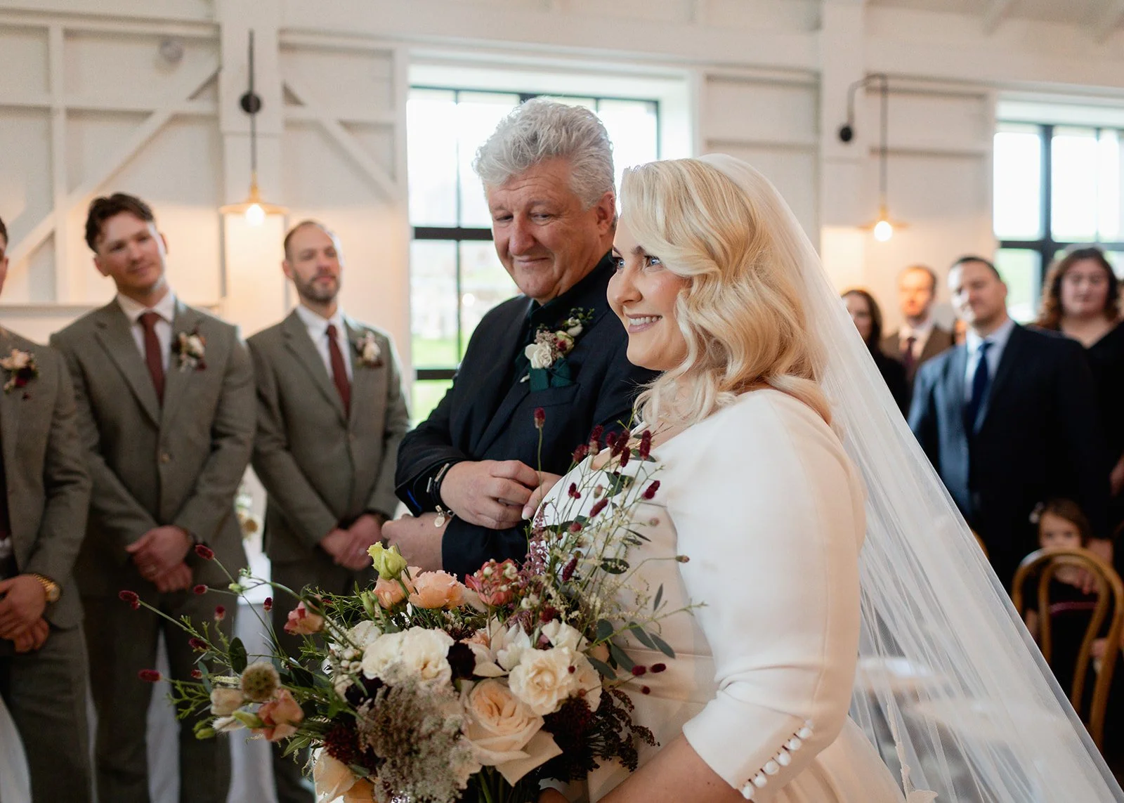 A bride and a man, likely her father, smiling and standing together during a wedding ceremony inside a bright, modern venue. The bride holds a large bouquet of flowers and wears a white wedding dress with a veil. The man wears a dark suit with a boutonniere. Guests in formal attire are visible in the background.