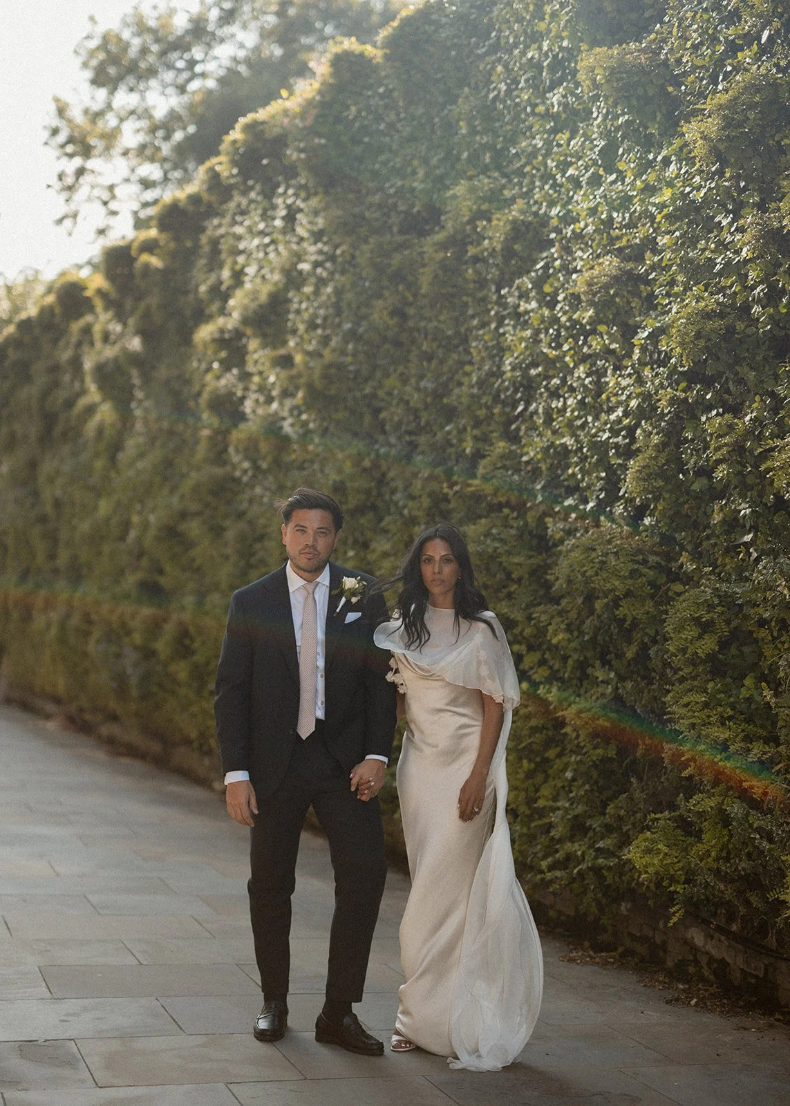 A couple dressed in wedding attire walking hand in hand outdoors on a paved path with a tall hedge of green foliage in the background, sunlight shining from the left side.