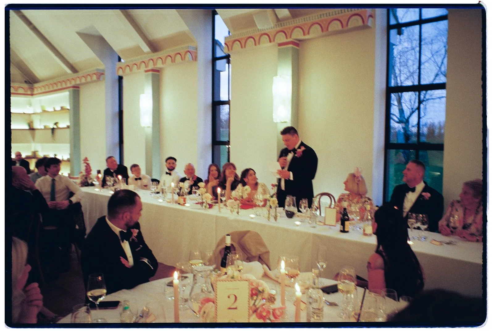 Wedding reception with a man giving a speech at a head table decorated with candles and flowers, surrounded by seated guests dressed in formal attire, inside a bright room with large windows and elegant architecture.