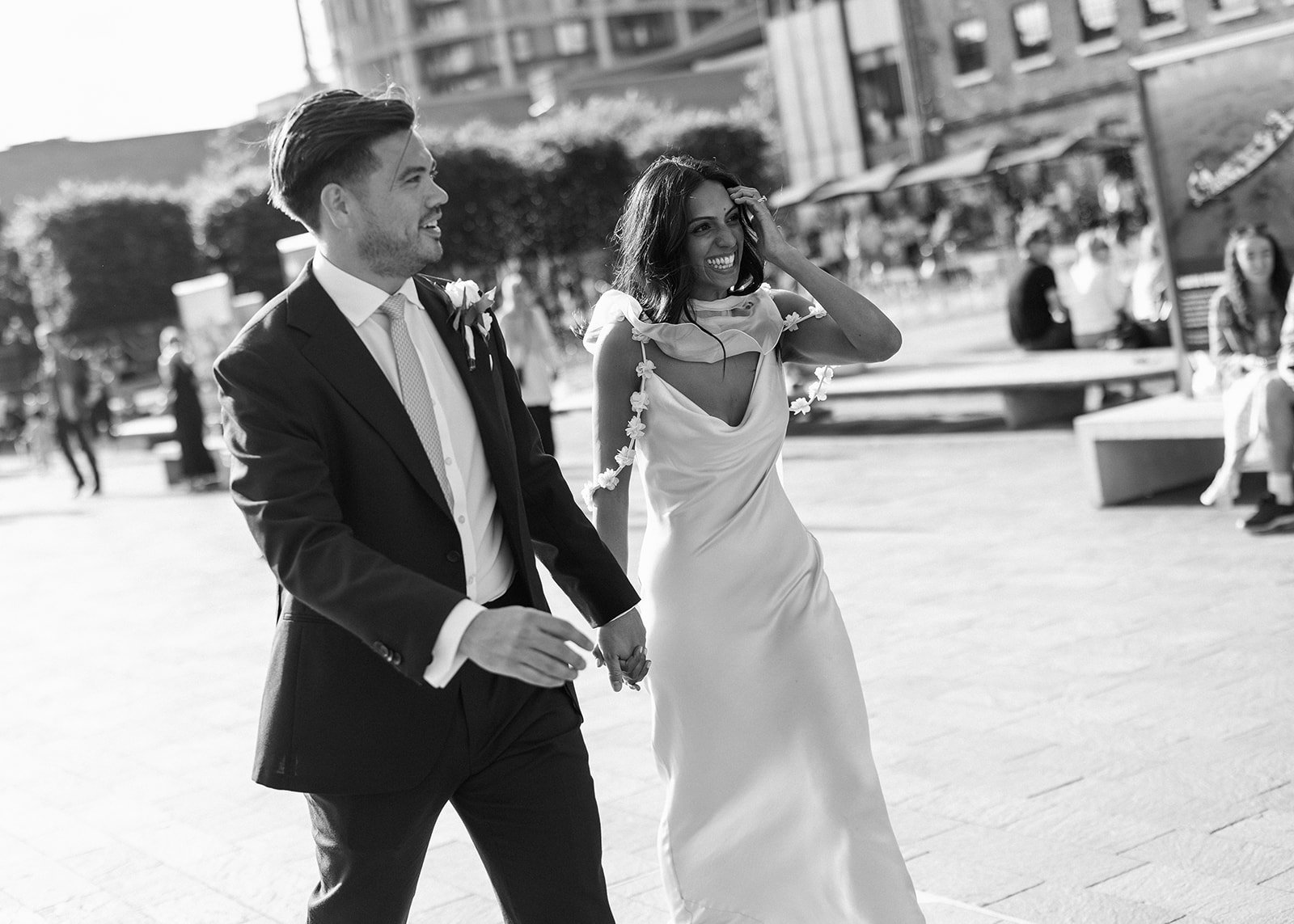 A black and white photo of a happily married couple, holding hands and walking outdoors, likely after their wedding ceremony. The man is dressed in a suit and tie, and the woman is in a wedding dress decorated with flowers, smiling broadly.