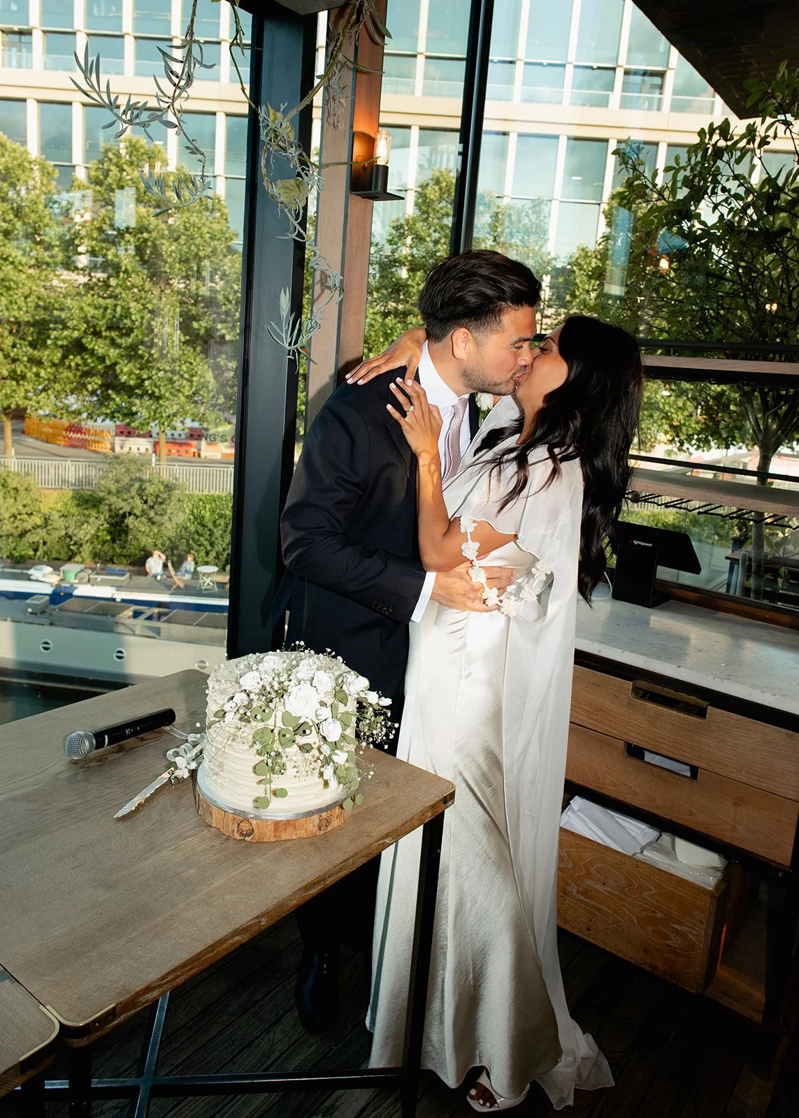 A couple in wedding attire sharing a kiss in a modern indoor location with large windows, a wedding cake decorated with white flowers on a wooden table, and a microphone nearby.