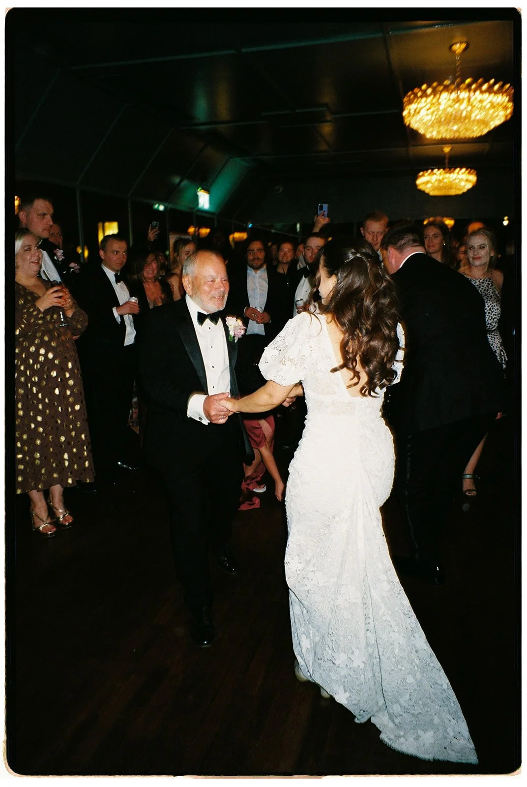 A bride and an older man dancing at a wedding reception surrounded by guests in a dimly lit venue with chandeliers.