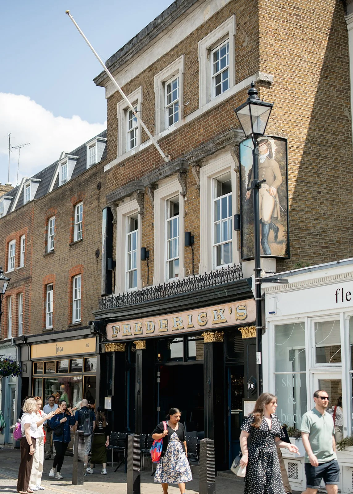 People walking on a busy street in front of Frederick's restaurant, with brick buildings, shop windows, and a lamppost with a decorative banner showing a woman in historical clothing.
