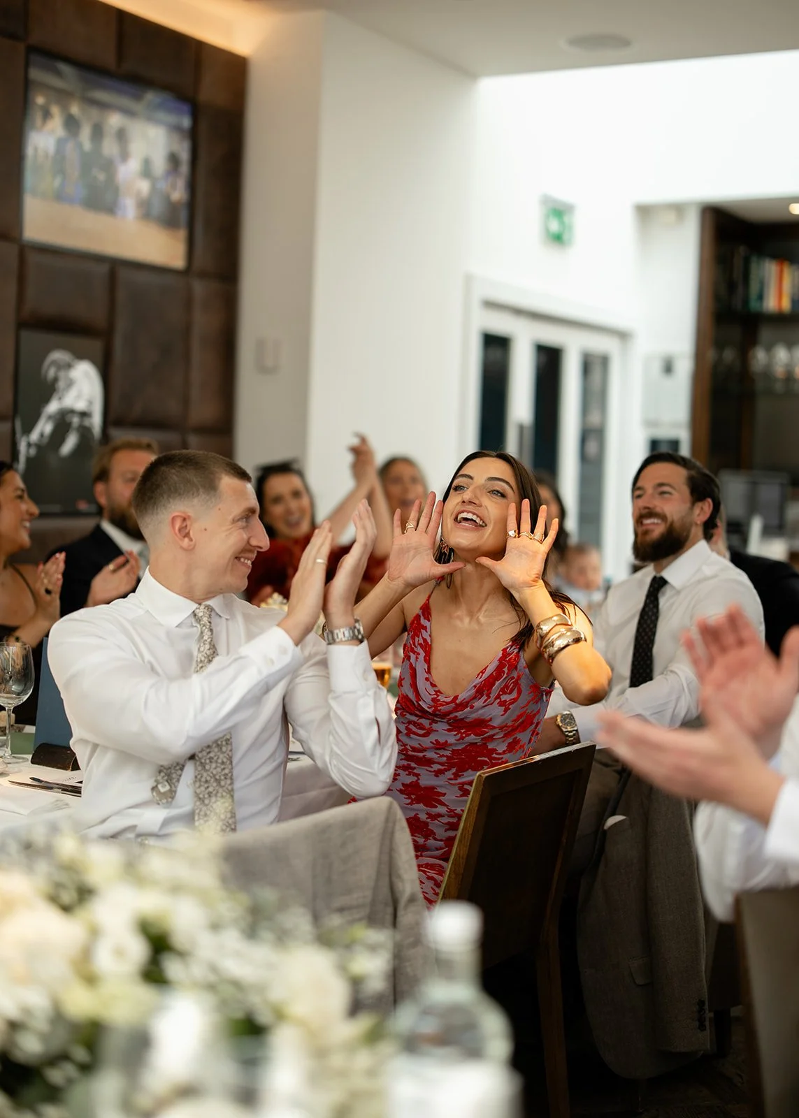 A woman in a red patterned dress is smiling and gesturing with her hands while sitting at a table during a celebration, surrounded by smiling people clapping and enjoying the event.