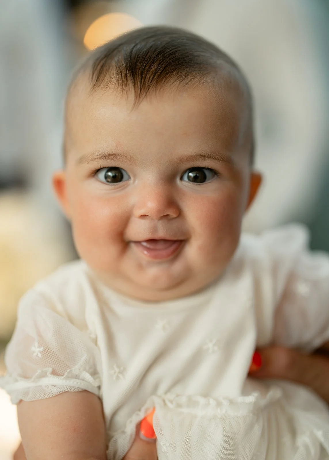 Close-up of a smiling baby with dark hair and big eyes, wearing a white dress with lace sleeves.