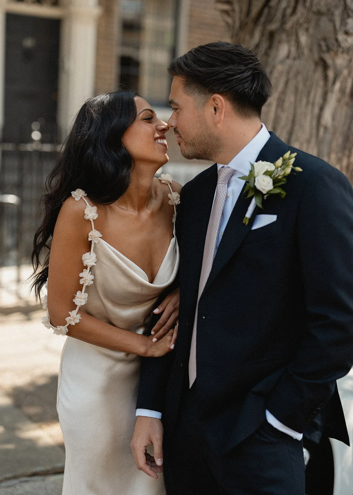 A bride and groom are close together, nose to nose, smiling, in an outdoor setting during a wedding.