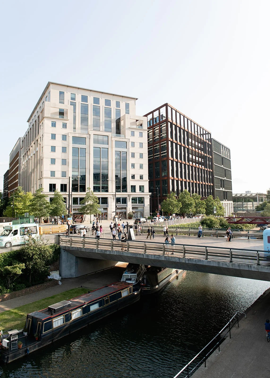Urban scene with modern office buildings, a busy street with people walking, cars, and a canal with a boat underneath a bridge.
