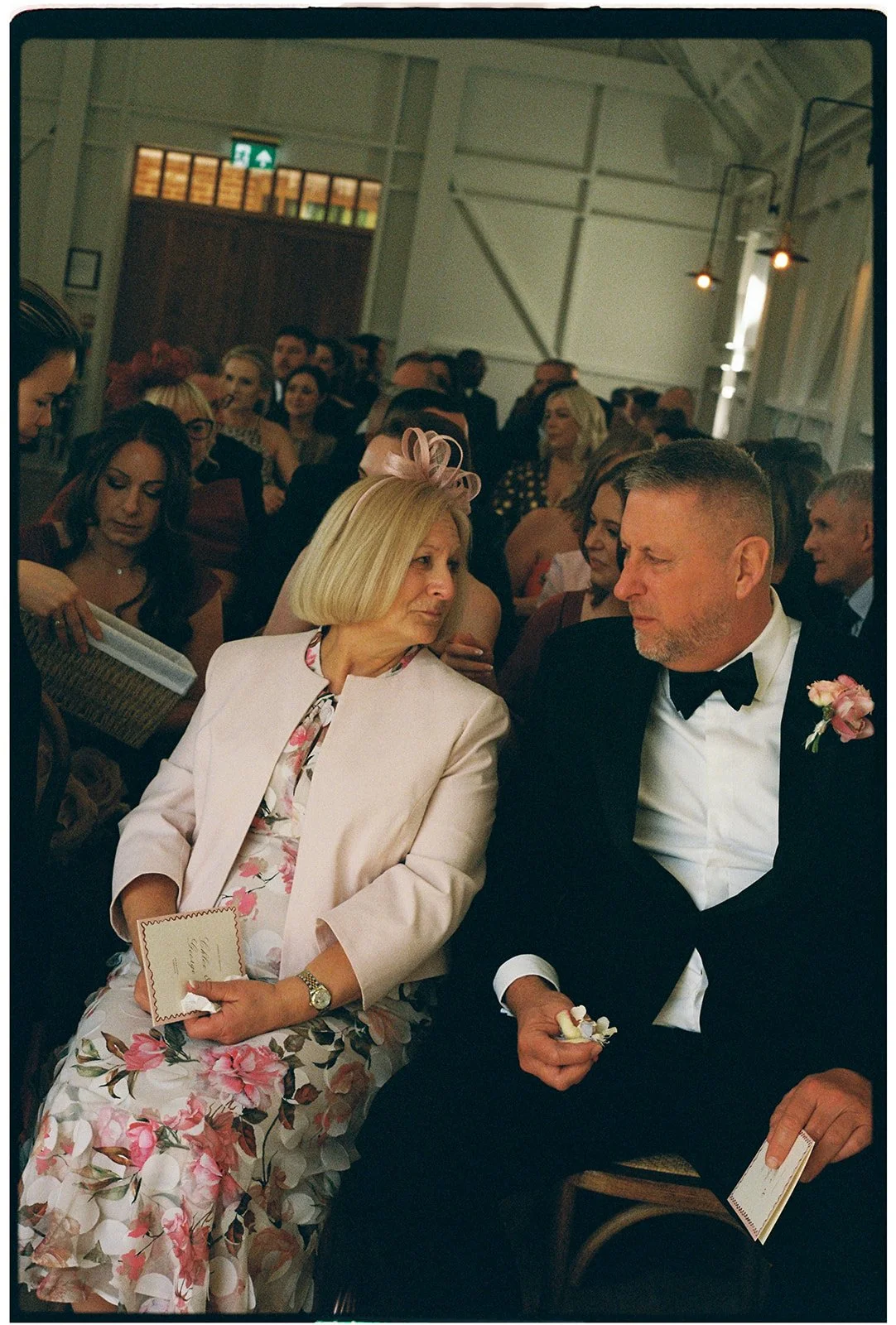 A wedding reception with guests seated in a decorated venue, including an older woman and a man in a tuxedo sitting close and conversing.