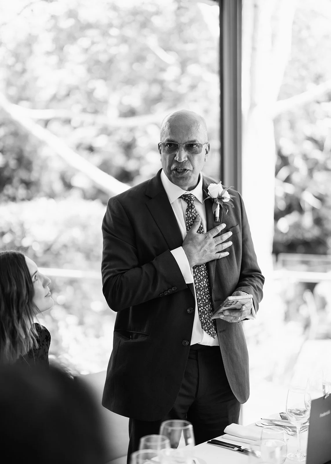 A man in a suit with a boutonniere speaks with his hand on his chest inside a building, possibly at a wedding or formal event.