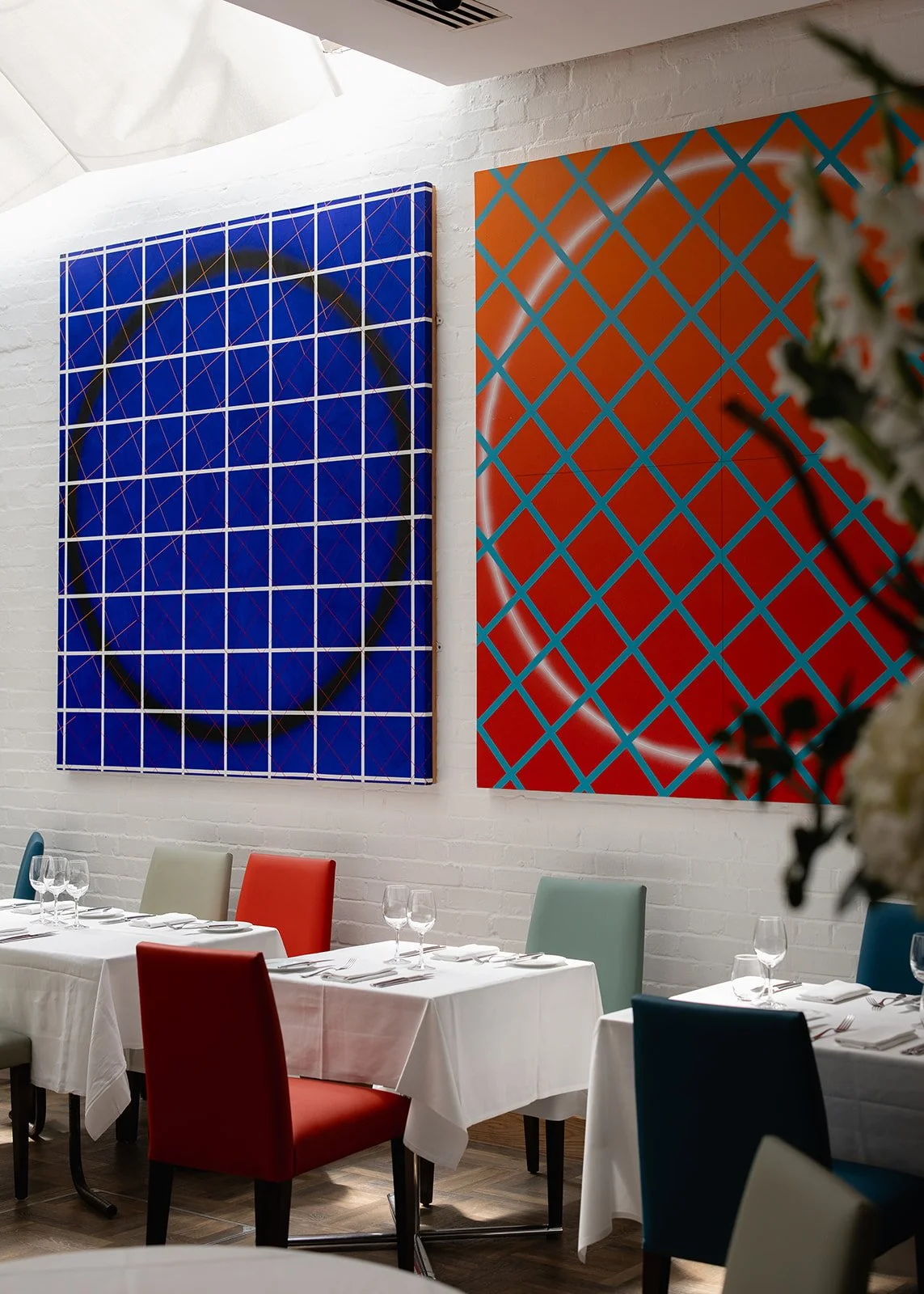 Artistic paintings hanging on a white brick wall in a modern dining area with tables set for a meal, including wine glasses, white tablecloths, and colorful chairs.