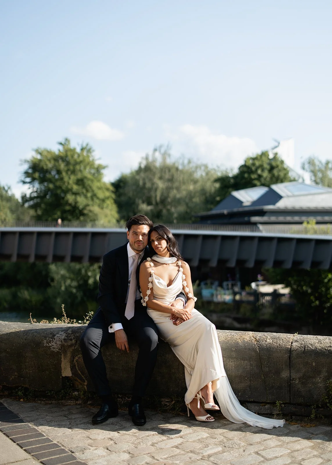 A man and woman sitting on a stone ledge outdoors, with the man wearing a black suit and tie and the woman wearing a white dress with floral details, both looking at the camera.