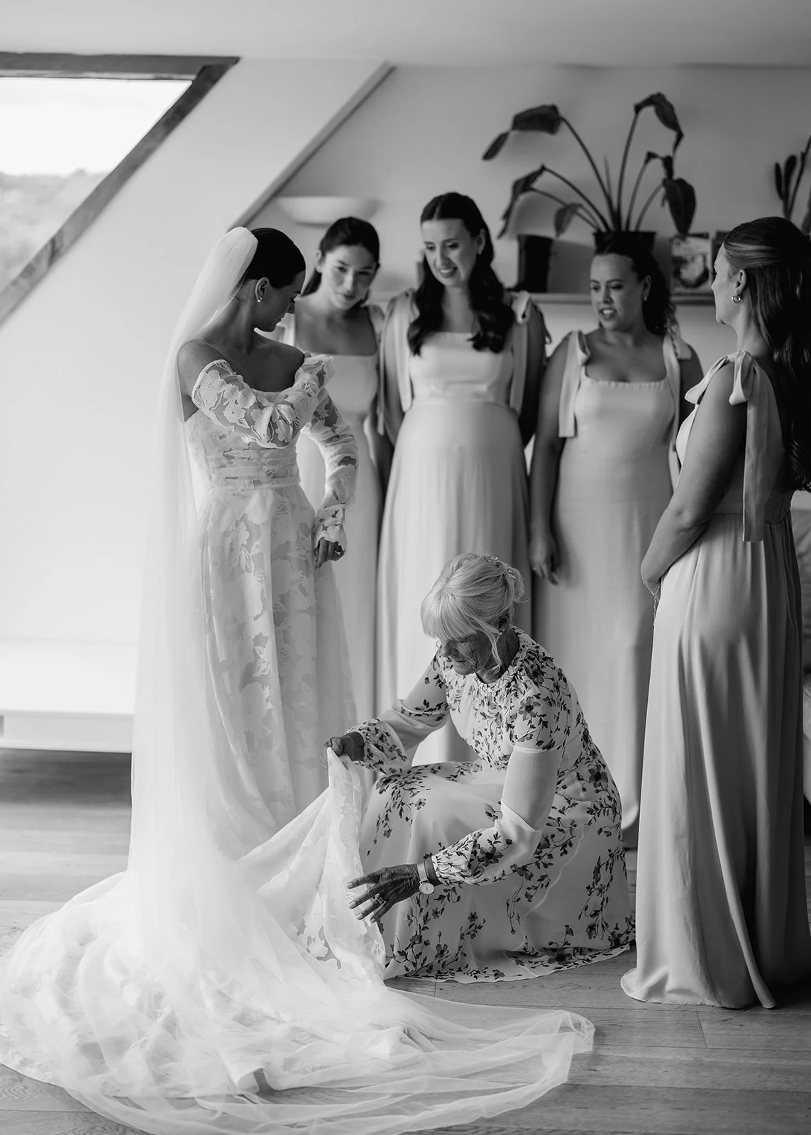 A bride in a wedding gown and veil is surrounded by five bridesmaids. An elderly woman is kneeling and adjusting the bride's train. Everyone is indoors, near a large window, in a room with plants.