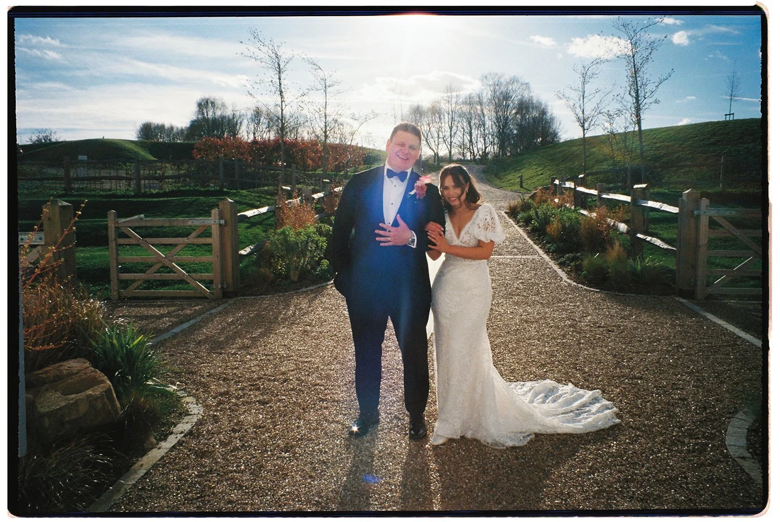 A bride and groom posing outdoors on a sunny day, with the bride wearing a white wedding dress and the groom in a tuxedo with a bow tie, standing on a gravel path surrounded by greenery and a rustic fence.