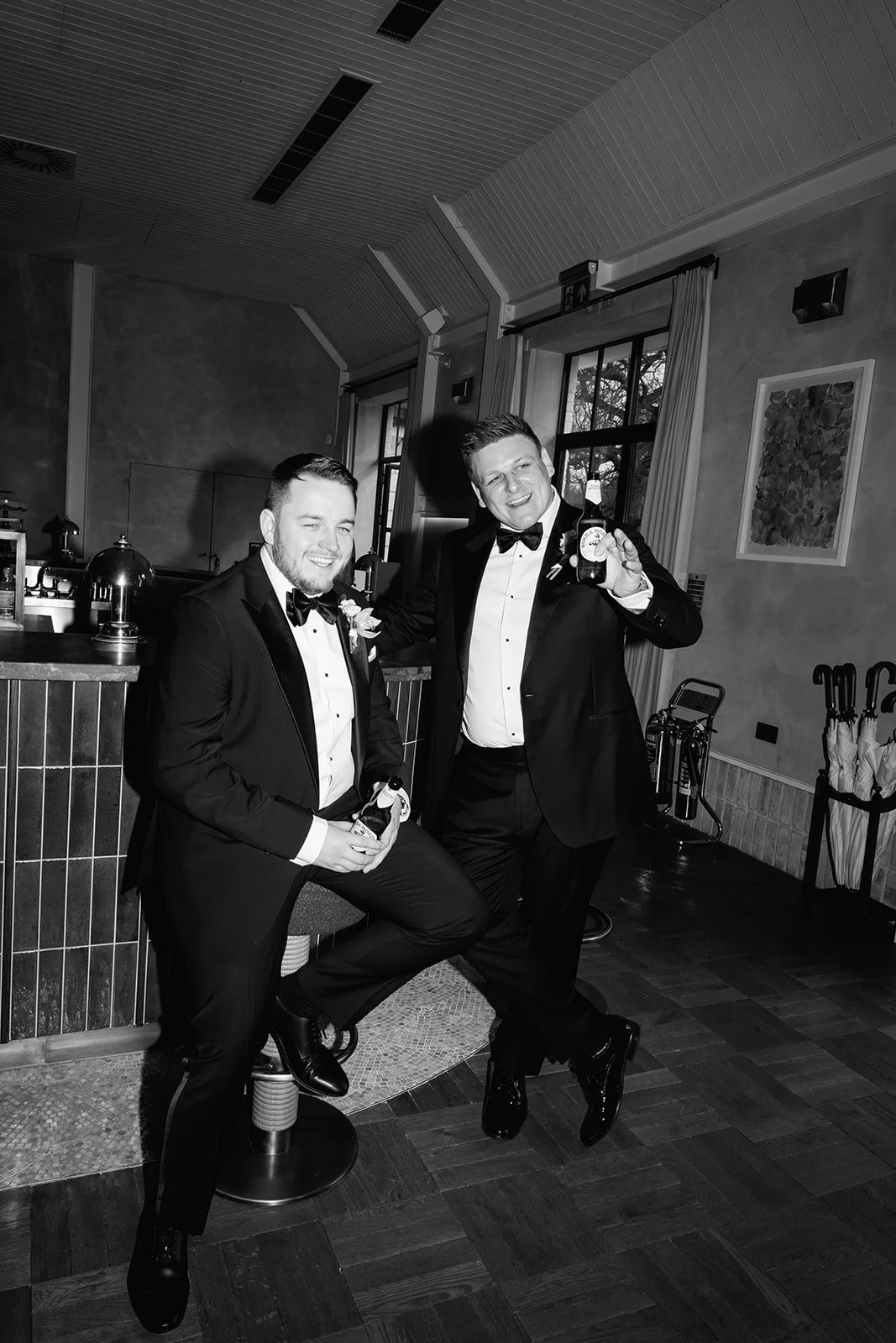 Two men in tuxedos celebrating at a wedding reception, holding drinks, smiling, and posing together indoors.
