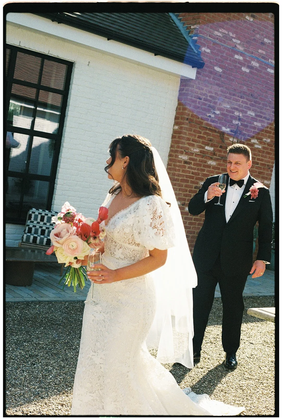 A bride holding a bouquet of pink and cream flowers and a champagne glass, with a groom in a black tuxedo and bow tie holding a glass of champagne, smiling, outdoors at a wedding reception near a brick wall and a white house.