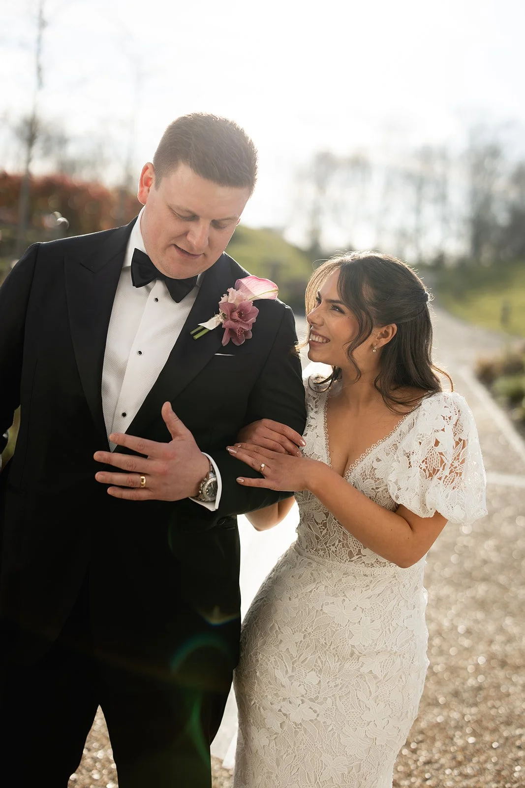 A bride and groom dressed in wedding attire, outdoors on a sunny day. The woman is smiling while touching the man's arm.